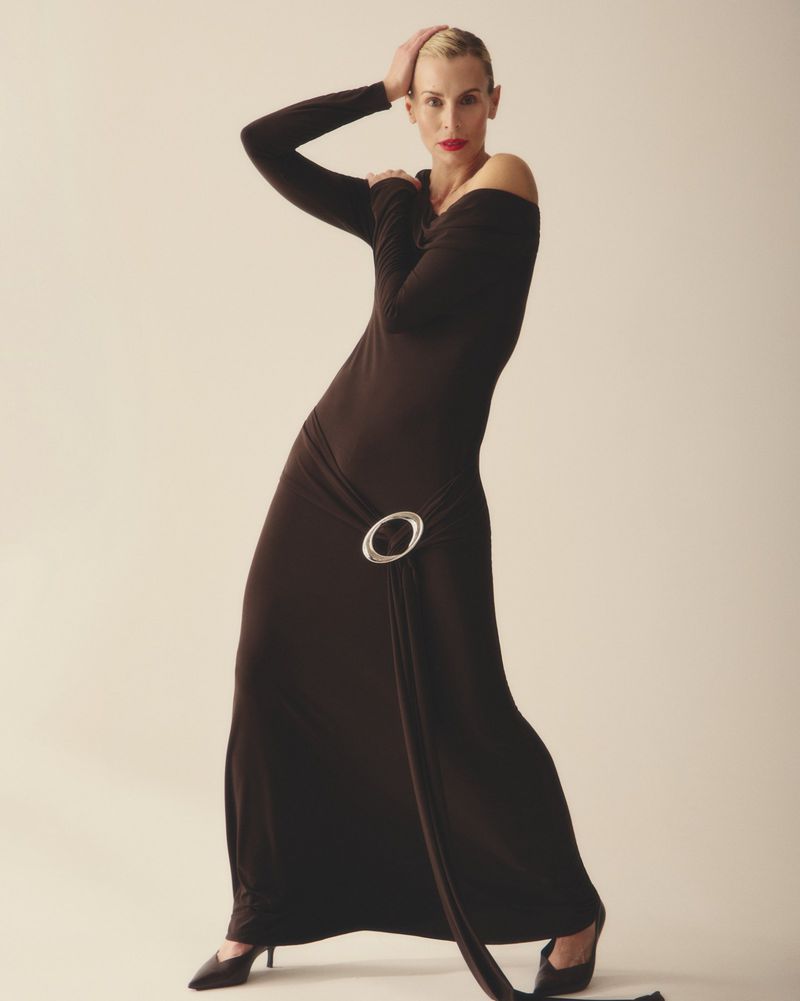 Model Niki Taylor wearing a long dark brown dress with a silver hip buckle against a seamless background