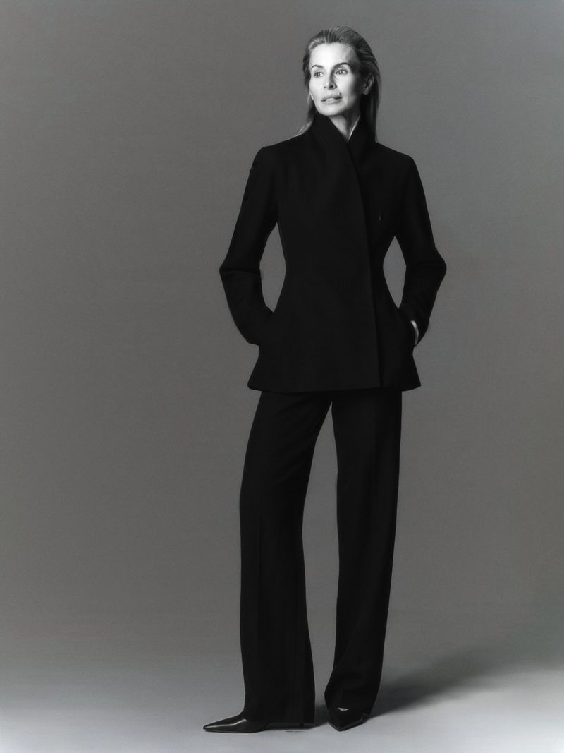 Portrait of a mature woman in a black tailored suit, standing in a photo studio setting, with a serious facial expression.
