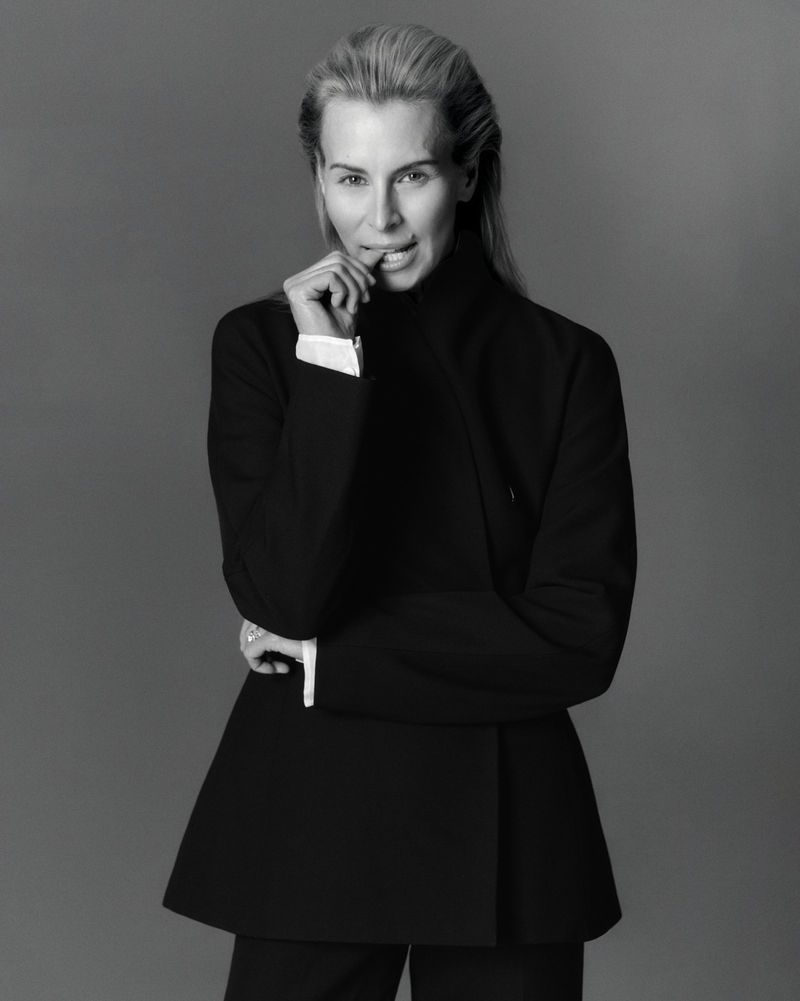 Elegant portrait of a blonde woman in a black suit against a gray backdrop, biting her finger in a thoughtful, poised manner.