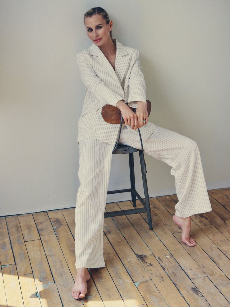 Woman in a striped pantsuit sitting on a stool. She has blonde hair and is smiling, studio lighting, hardwood floors, indoor fashion shoot.