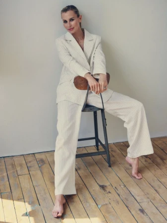 Woman in a striped pantsuit sitting on a stool. She has blonde hair and is smiling, studio lighting, hardwood floors, indoor fashion shoot.