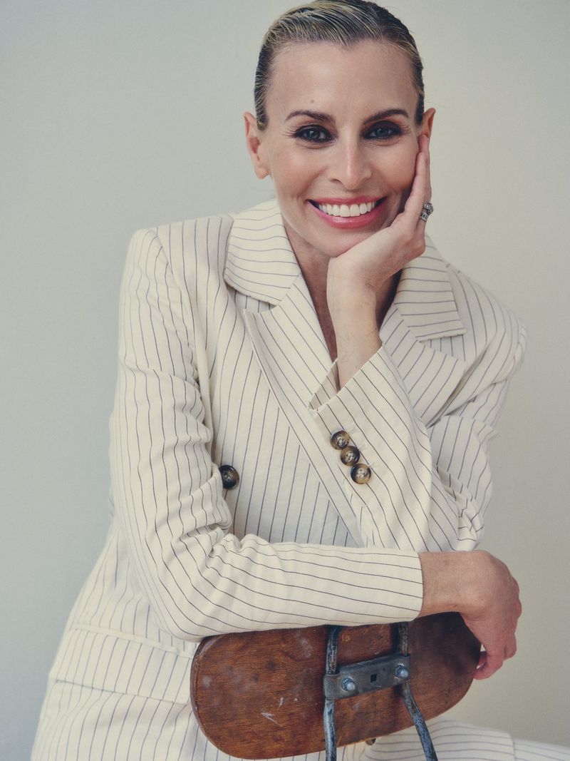 Portrait of a smiling woman in a striped suit, posing in a studio with a neutral background while seated on a wooden chair.