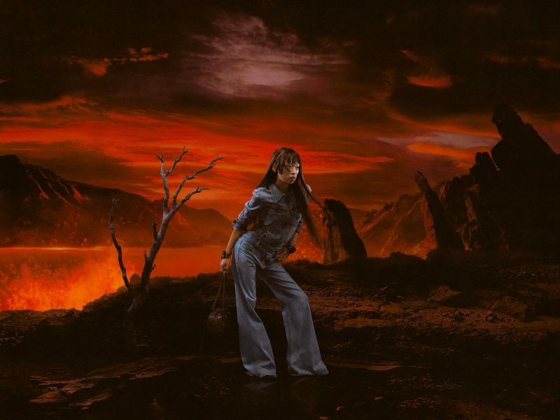 Woman poses in denim outfit against a surreal landscape with a fiery sky and jagged terrain, holding a drawstring bag.