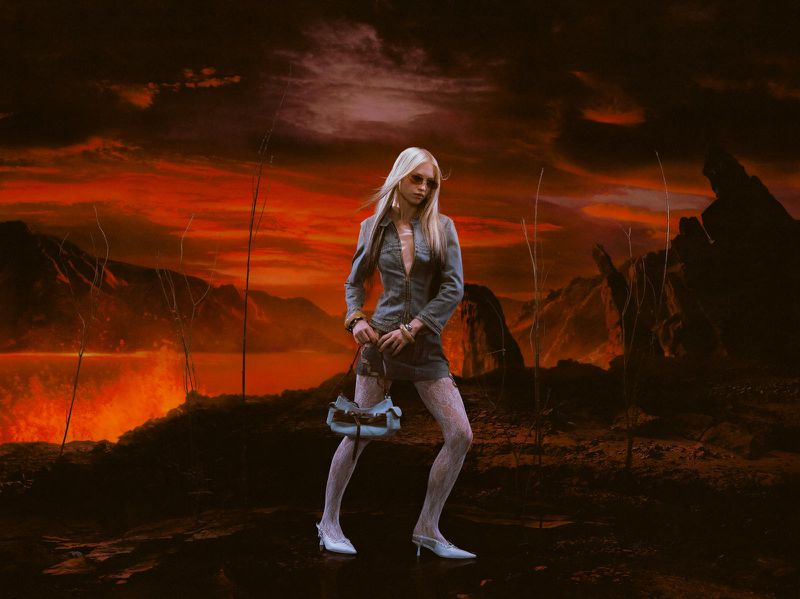 Fashionable blonde woman in grey suit, white fishnet stockings and heels poses with a handbag against a dramatic volcanic landscape.