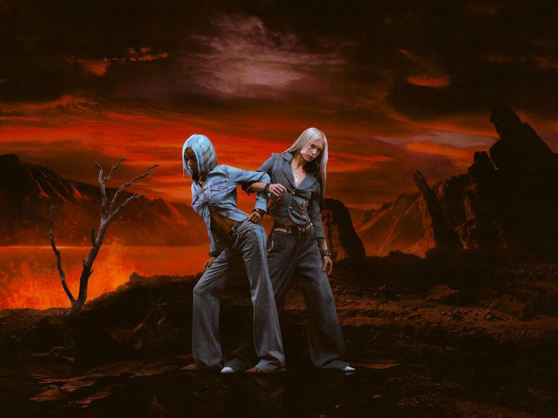 Two women posing in denim outfits against a backdrop of a red, mountainous landscape under a dramatic sky with a lava lake.