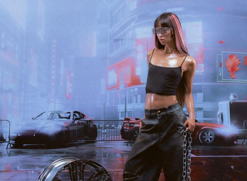 Adult female model with pink-streaked hair in a futuristic urban scene with cars, wearing a black crop top, pants, and silver chain.