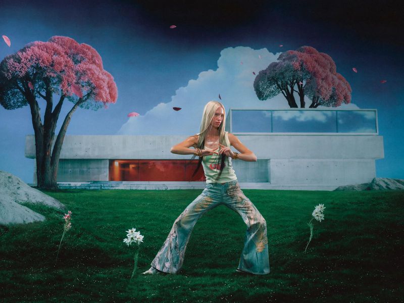 Woman strikes a pose in a vibrant outdoor setting with flowering trees and a modern building, blending fashion with nature's surreal charm.