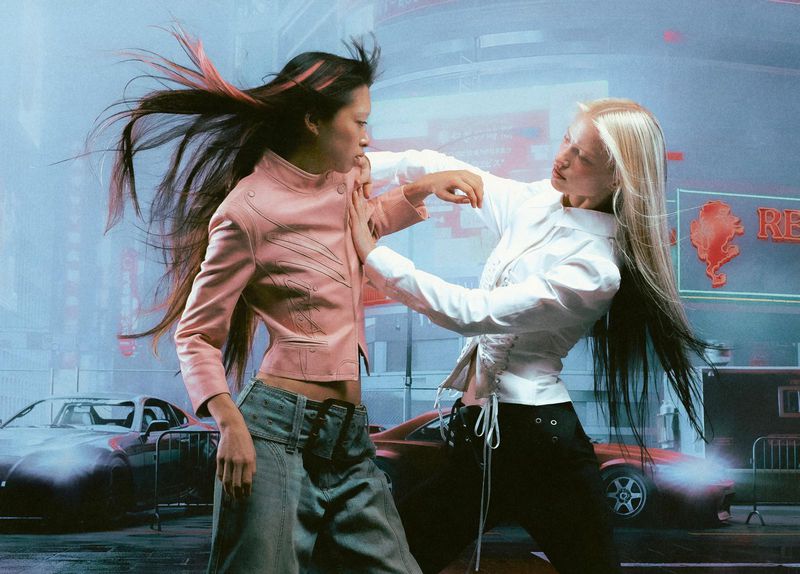 Two women face each other in urban scene, wearing stylish jackets and pants, neon lights and cars in background, fashion forward.