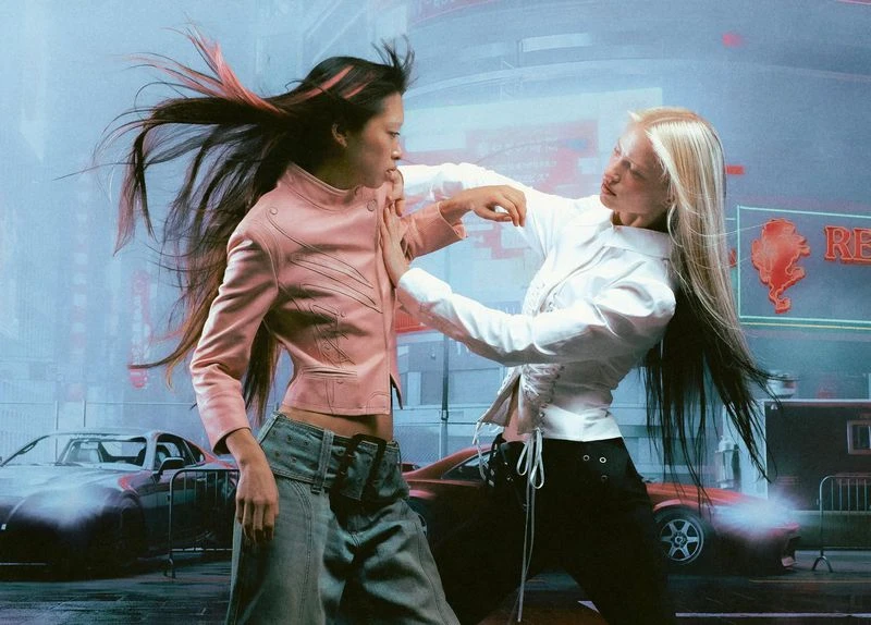 Two women face each other in urban scene, wearing stylish jackets and pants, neon lights and cars in background, fashion forward.