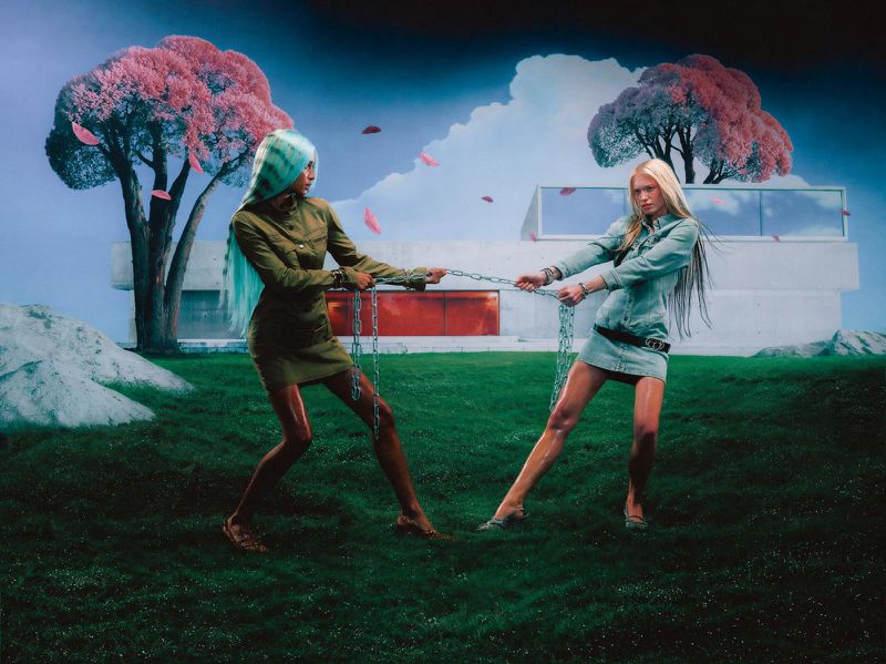 Two fashion models in dresses pull on a chain in a grassy yard with pink trees and a modern house with a red accent wall in the background.