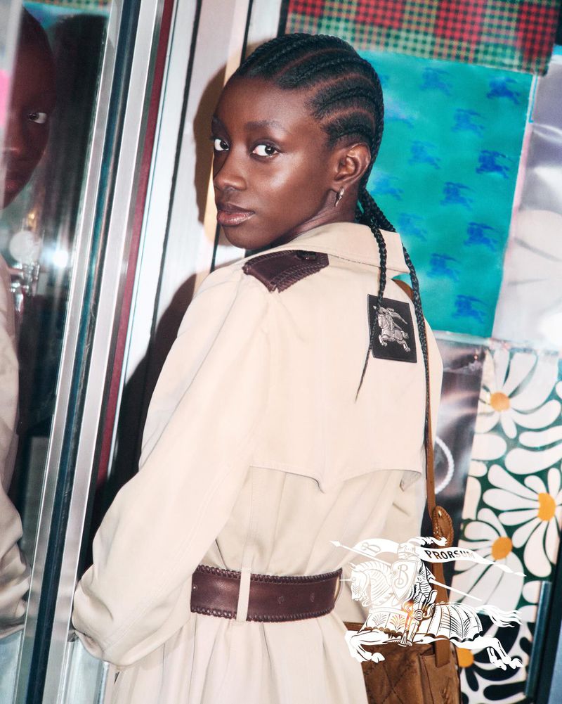 Fashion shot of a Black woman in a tan trench coat with a brown belt, braided hair, and silver earrings against a colorful patterned backdrop.