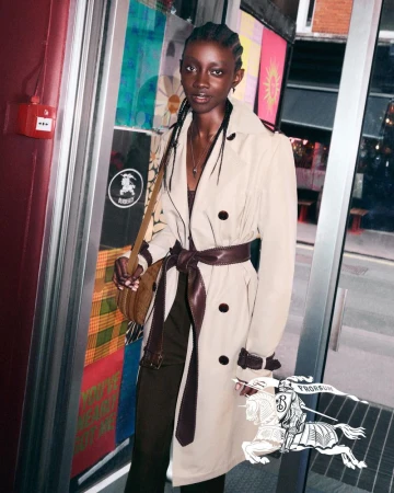 An adult woman wearing a belted trench coat stands in a doorway. She carries a shoulder bag in an urban environment with colorful backgrounds.
