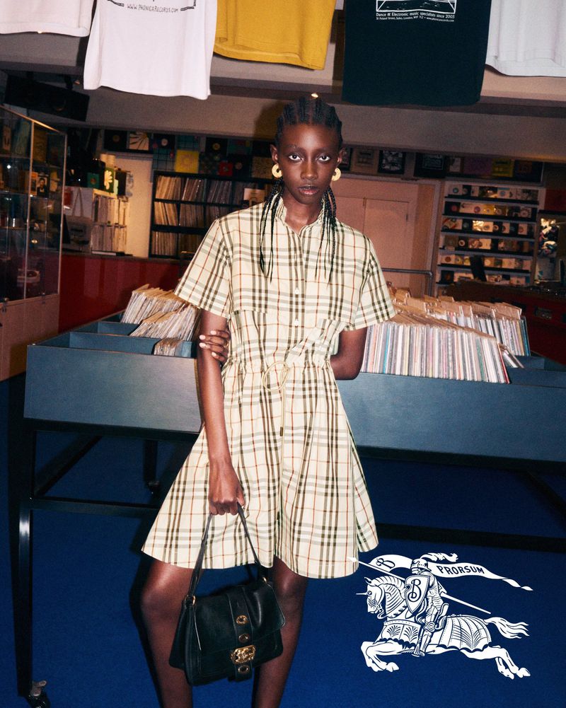 A dark-skinned woman poses in a tan and plaid dress, holding a black purse in a music store with vinyl records in the background.