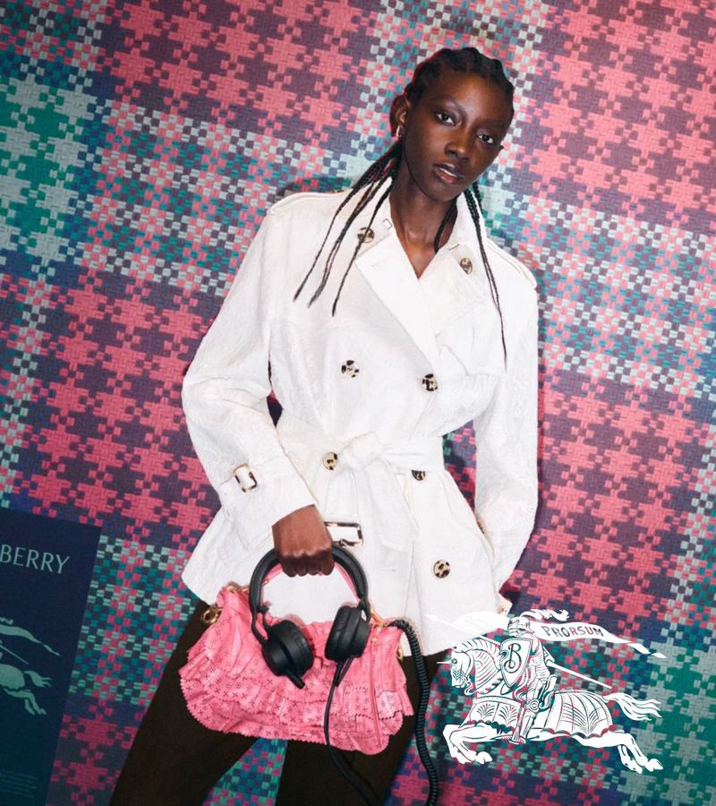 Model in white trench coat holding a pink handbag against a pink plaid background