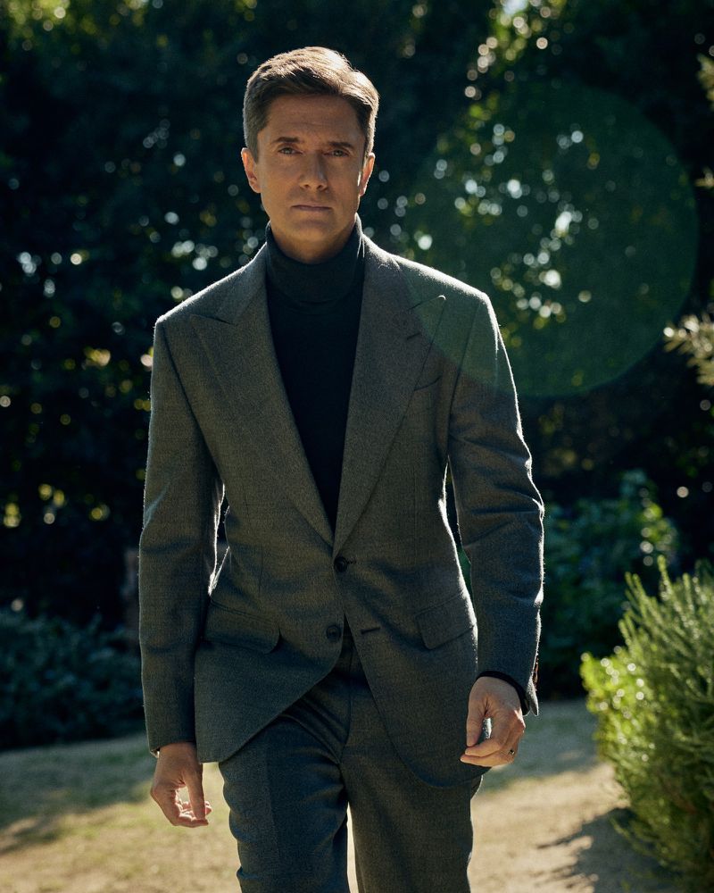 A man in a grey suit walks confidently outside on a sunny day. The background includes green foliage and bokeh, creating a natural setting.