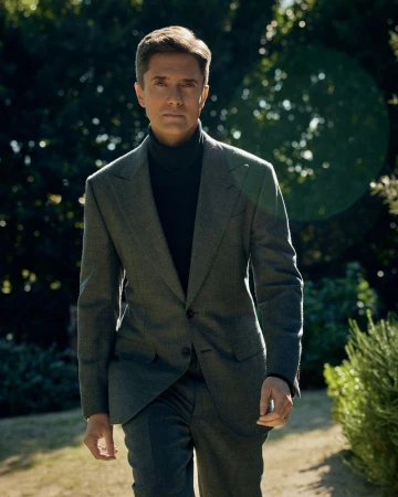 A man in a grey suit walks confidently outside on a sunny day. The background includes green foliage and bokeh, creating a natural setting.