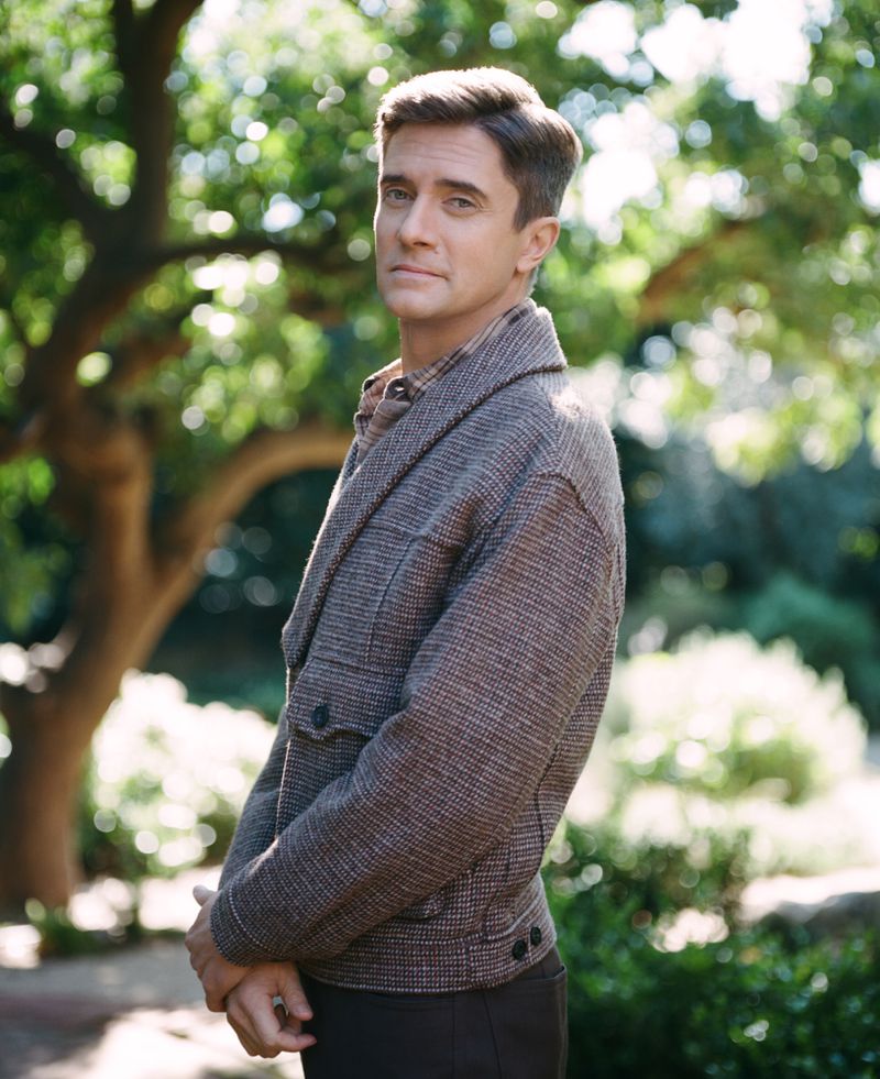 Adult male poses in a brown tweed jacket outdoors with blurred green trees and foliage providing a natural, sunny backdrop.