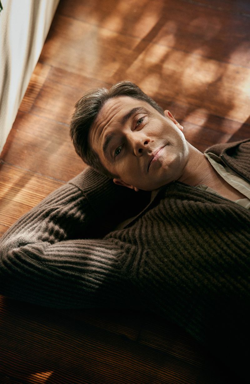 Portrait of an adult male lying on a wooden surface, wearing a brown cardigan, with natural light and shadows playing across the scene.