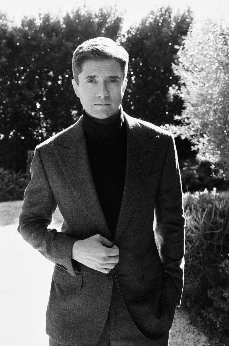 Portrait of Topher Grace wearing a dark blazer and turtleneck sweater outdoors against a garden background.