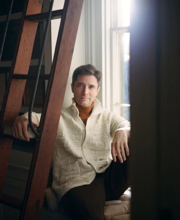 Adult male in white cardigan poses near window with natural light and a ladder inside a home interior, creating an editorial portrait.