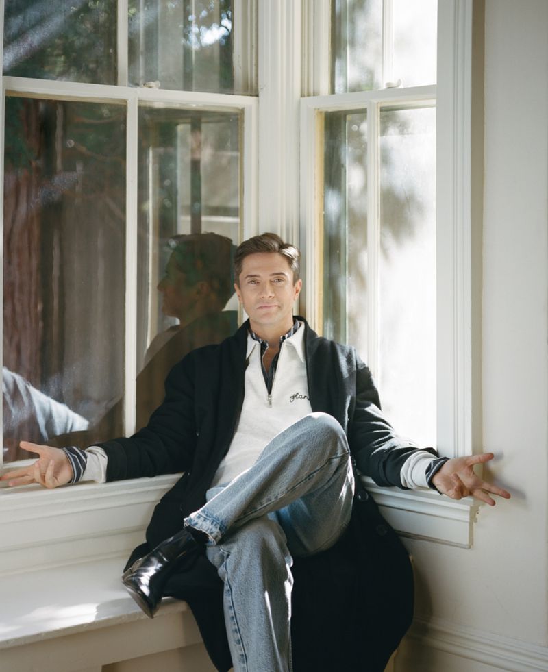 Adult male poses indoors near a window wearing a white collared shirt, light-wash denim jeans, and a long black jacket.