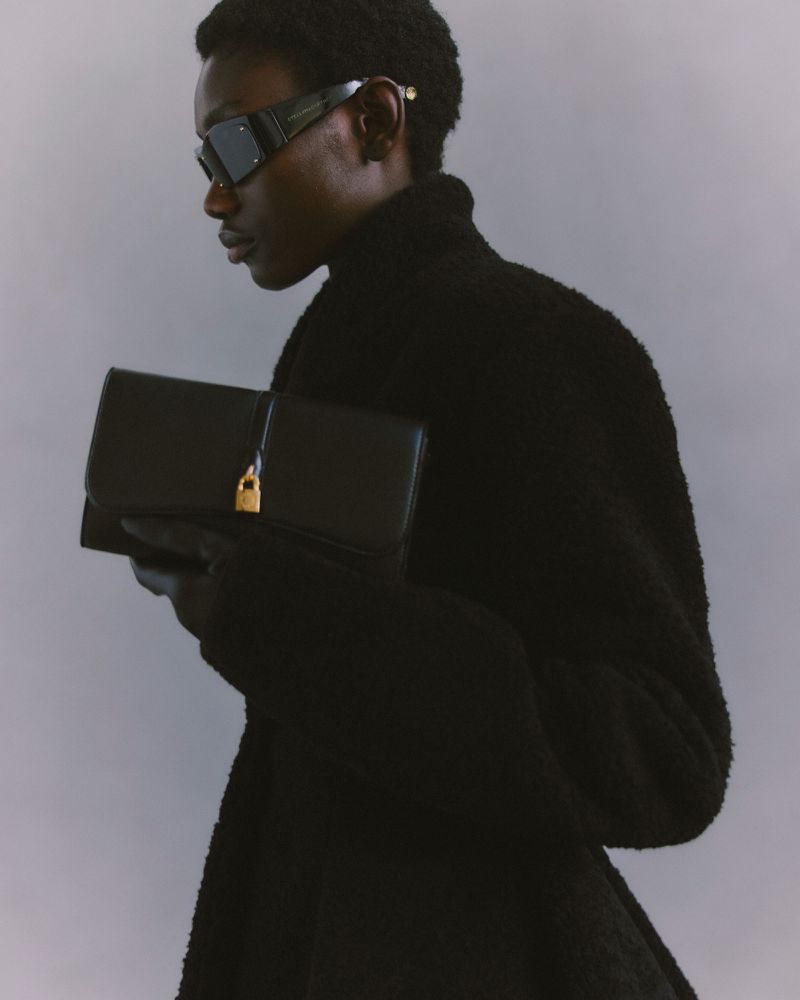 Stylish Black adult in a black coat and sunglasses, holding a black clutch bag with a gold lock, against a grey backdrop. 