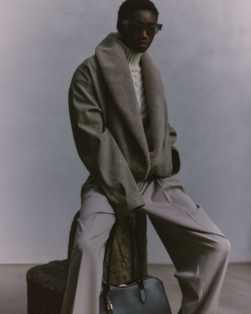 An adult male in a grey coat and dark sunglasses sits on a textured stool next to a black leather bag, with a light grey studio background.