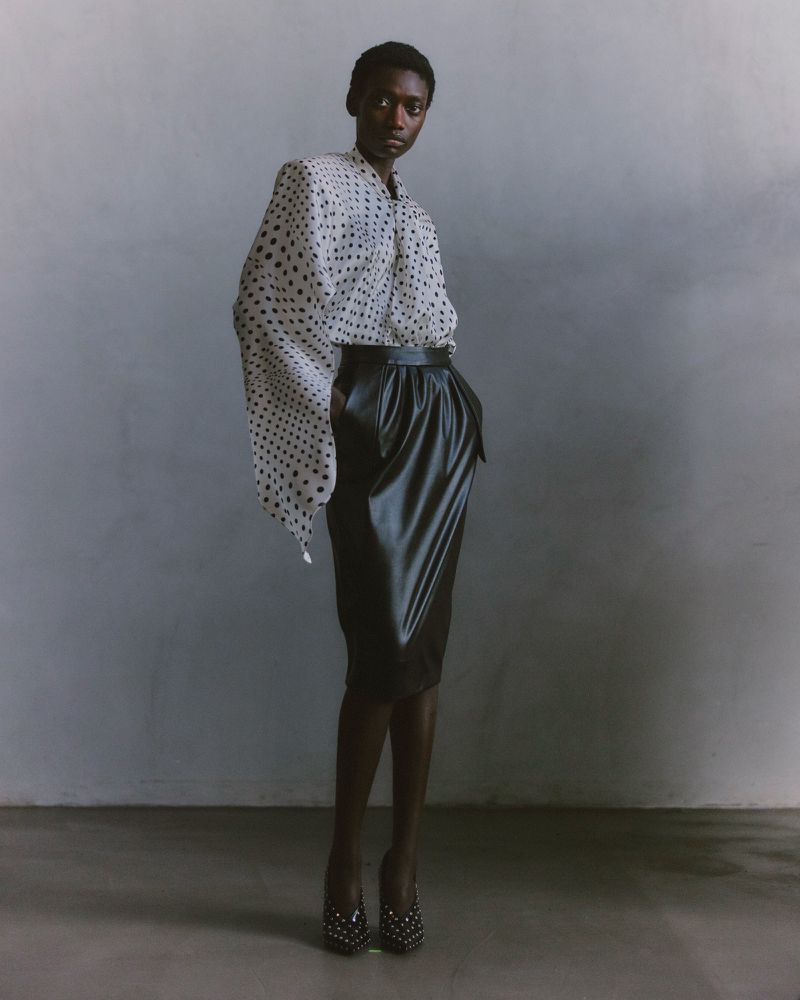 A striking full shot captures a Black female model in a polka dot blouse and black skirt, with a neutral background and studio lighting.