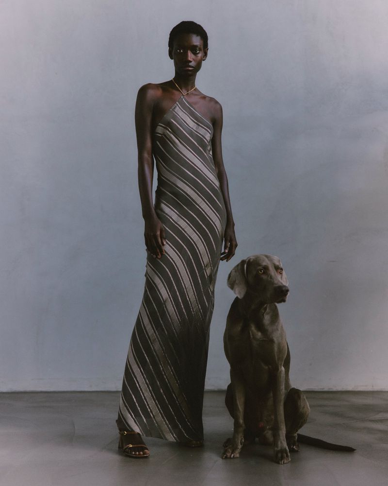 A Black woman in a striped dress poses with a grey Weimaraner dog in a studio setting with a simple background and neutral tones.