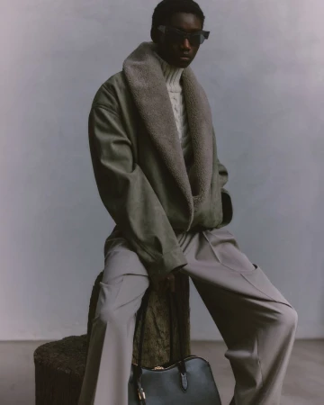 Model wearing a shearling-collar jacket and sunglasses, holding a black handbag against a grey background.