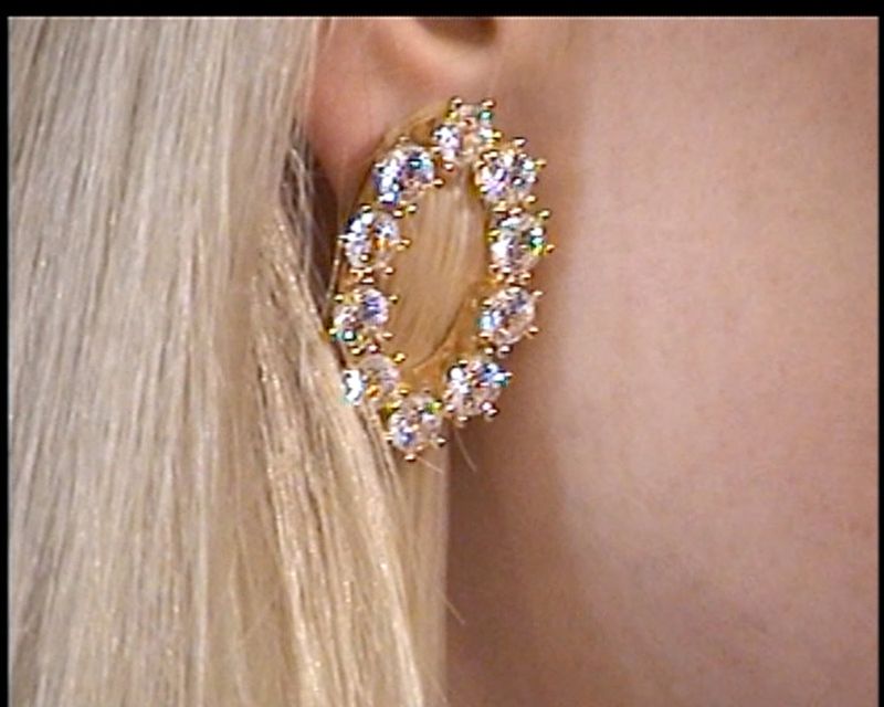 Close-up of a blonde adult female with a large gold oval earring encrusted with clear gemstones, showcased against a neutral skin tone.