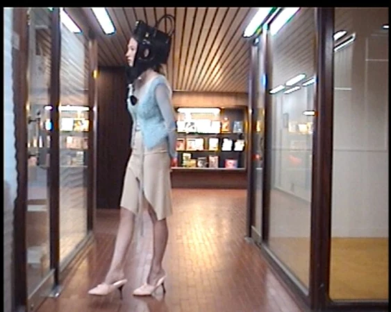 Woman in a light blue top and khaki skirt stands in a hallway with display windows, wearing a black headgear device.
