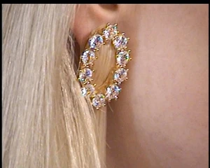 Close-up of a gold earring encrusted with round white crystals worn on an earlobe next to blonde hair.