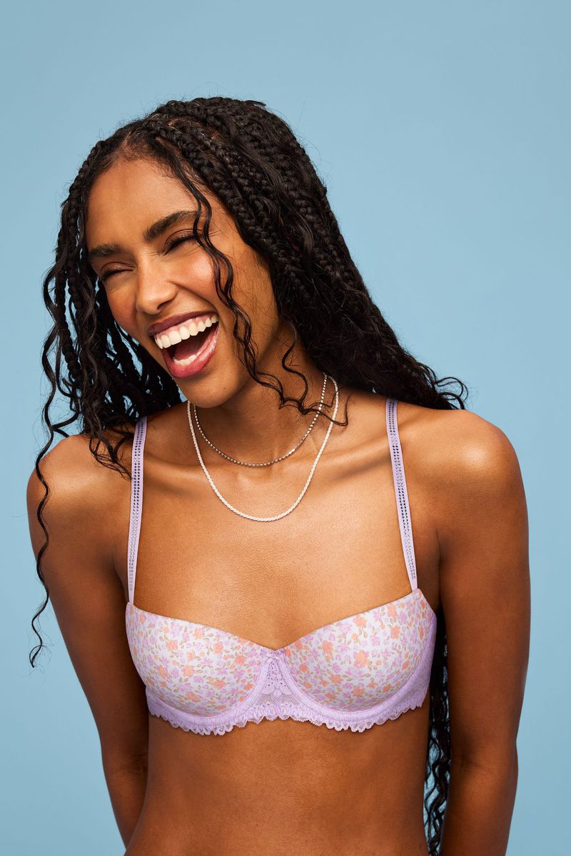 Smiling dark-skinned woman with braided hair wears a lilac floral bra against a blue backdrop, showcasing natural beauty and undergarment fashion.