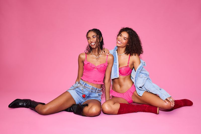 Two beautiful Black women pose in pink lingerie and denim shorts against a bright pink background in a fashion studio setting.