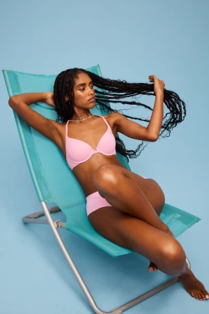 A woman in pink swimwear relaxes on a light blue lounge chair against a matching backdrop, showcasing fashion and leisure.