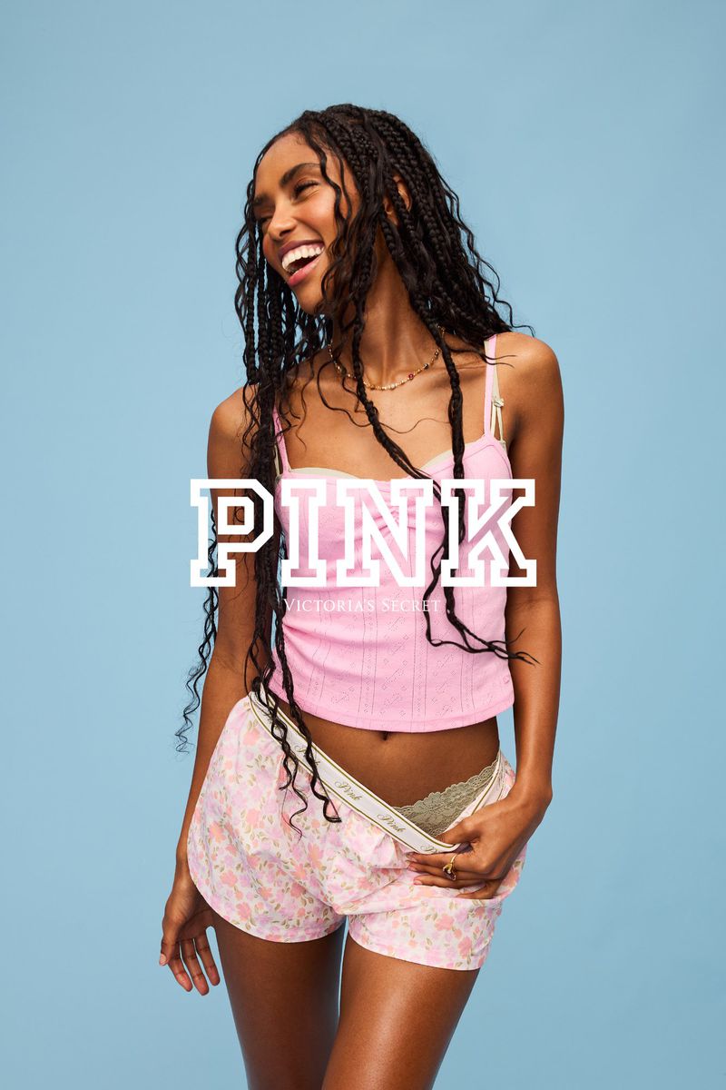 A smiling young woman wearing a pink top and floral shorts poses in a studio environment against a light blue backdrop.