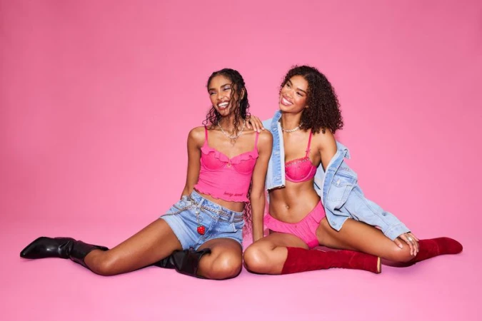 Two female models posing seated against a solid pink backdrop wearing pink lingerie and denim clothing for Victoria's Secret.