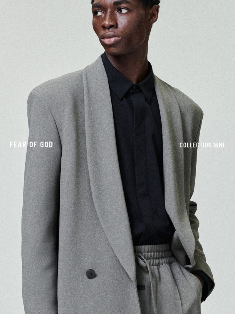 Fashion shot of a Black male model in a gray double-breasted blazer and matching pants, against a neutral background, looking at the camera.