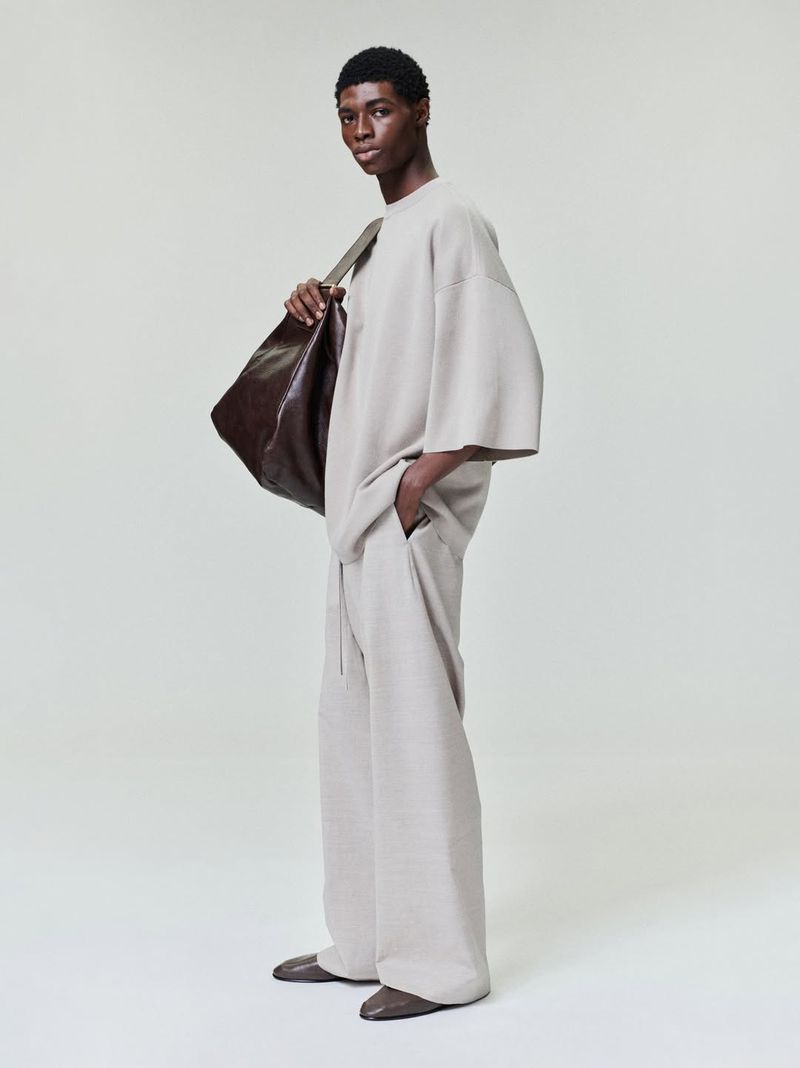 Studio shot of a Black male model in an oversized beige outfit with a brown leather bag, posing against a white backdrop.