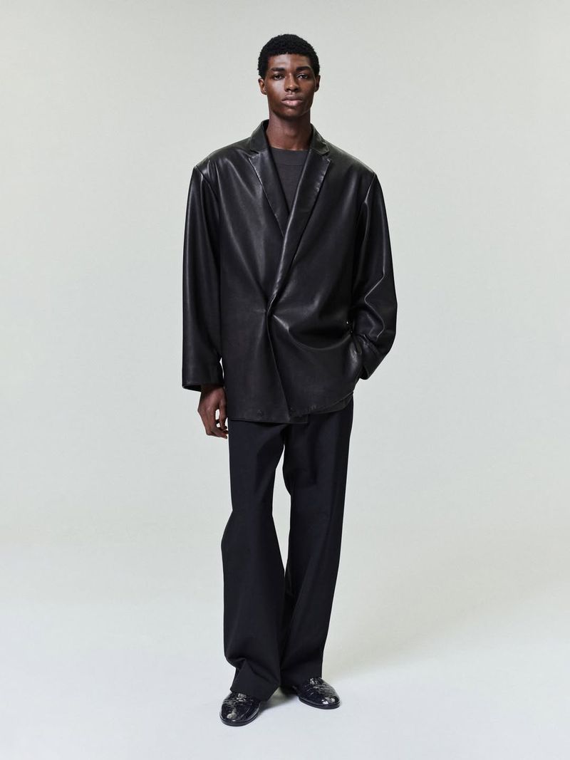 A full shot of a Black male model in a grey t-shirt, oversized leather blazer, and black trousers standing in a neutral studio setting.