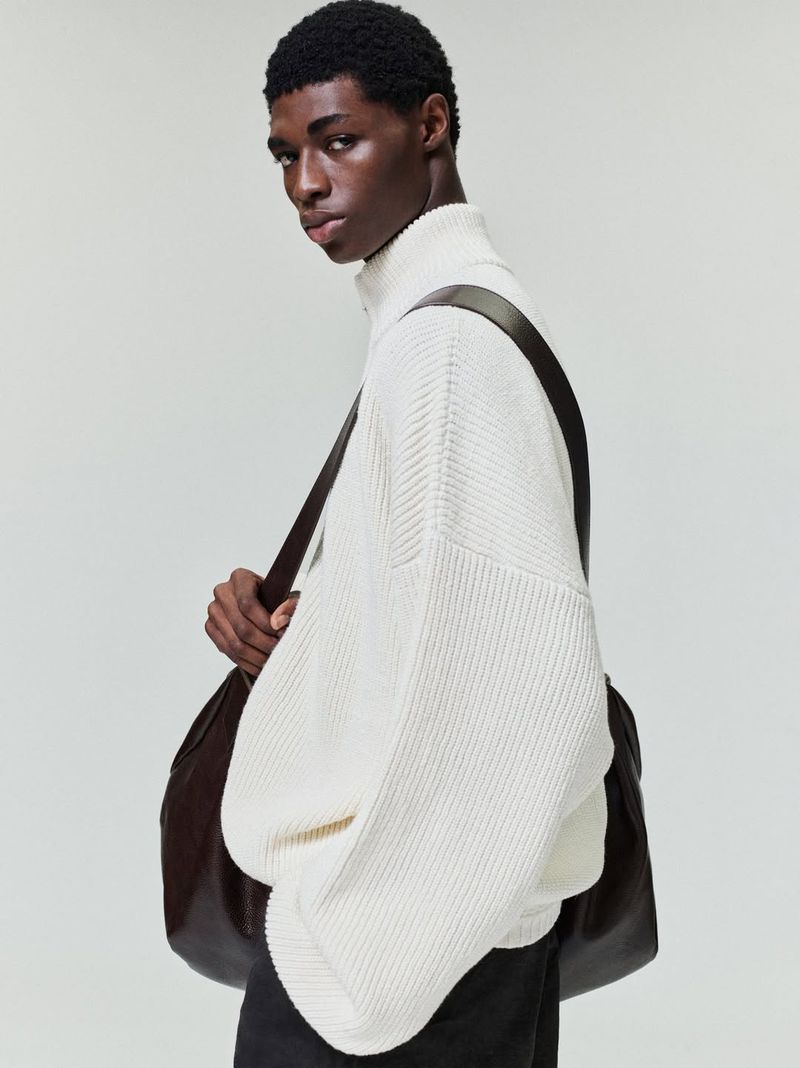 A young Black male model wearing a white sweater and carrying a dark leather bag poses in a studio with a neutral background.