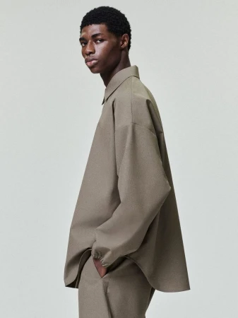 A male fashion model in a loose-fitting beige two-piece suit poses in a studio with soft, diffused lighting on a neutral background.