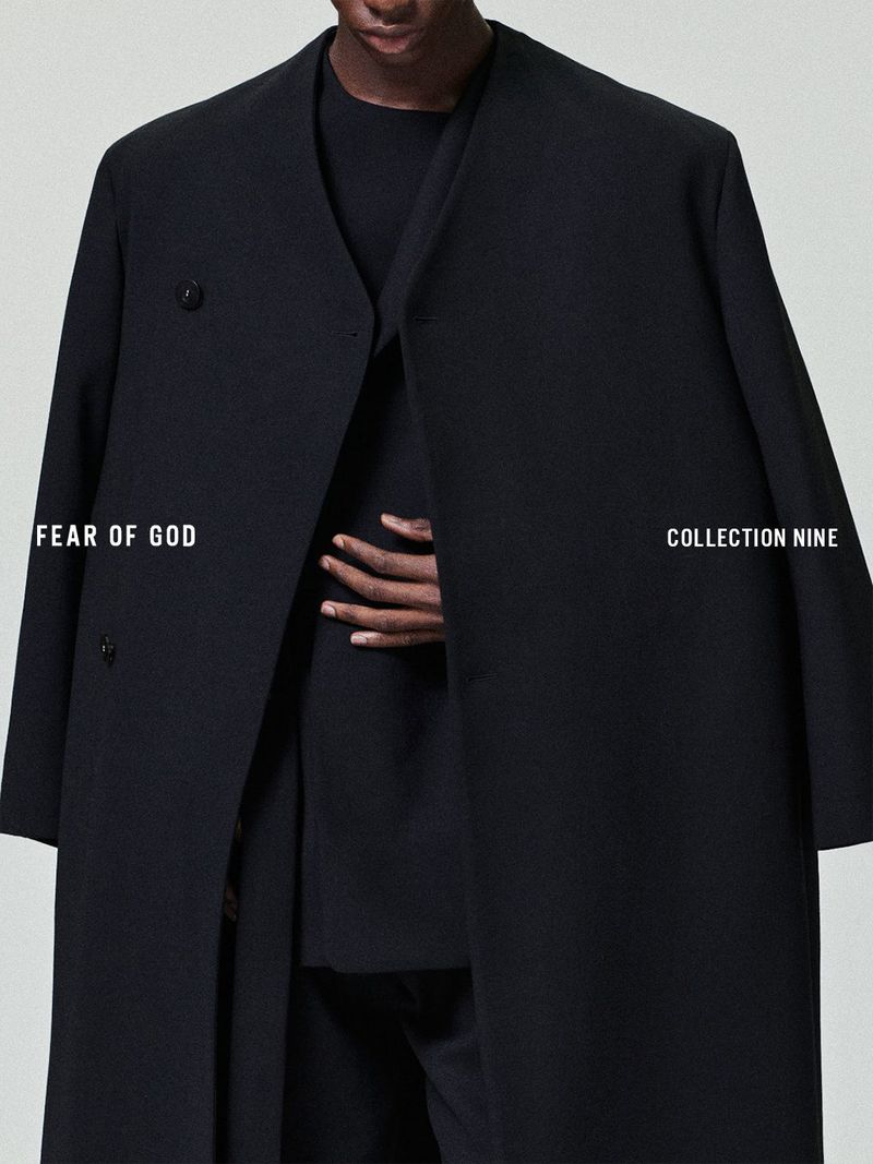 Fashion shot of a black coat on an adult male indoors. The coat is worn over a black outfit. Text overlay includes the brand and collection.