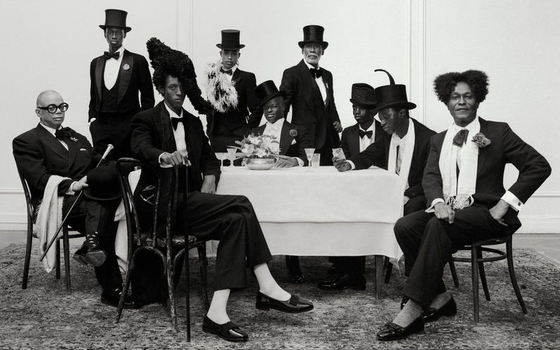 Group of eight African American men in formal wear, including tuxedos and top hats, gather around a table in an indoor setting.
