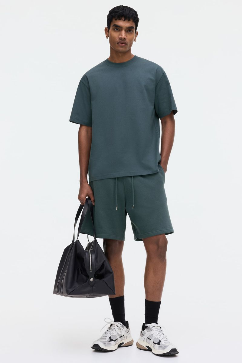 Man in a blue-green t-shirt and shorts stands in a white studio holding a black leather bag, wearing running shoes and black socks.