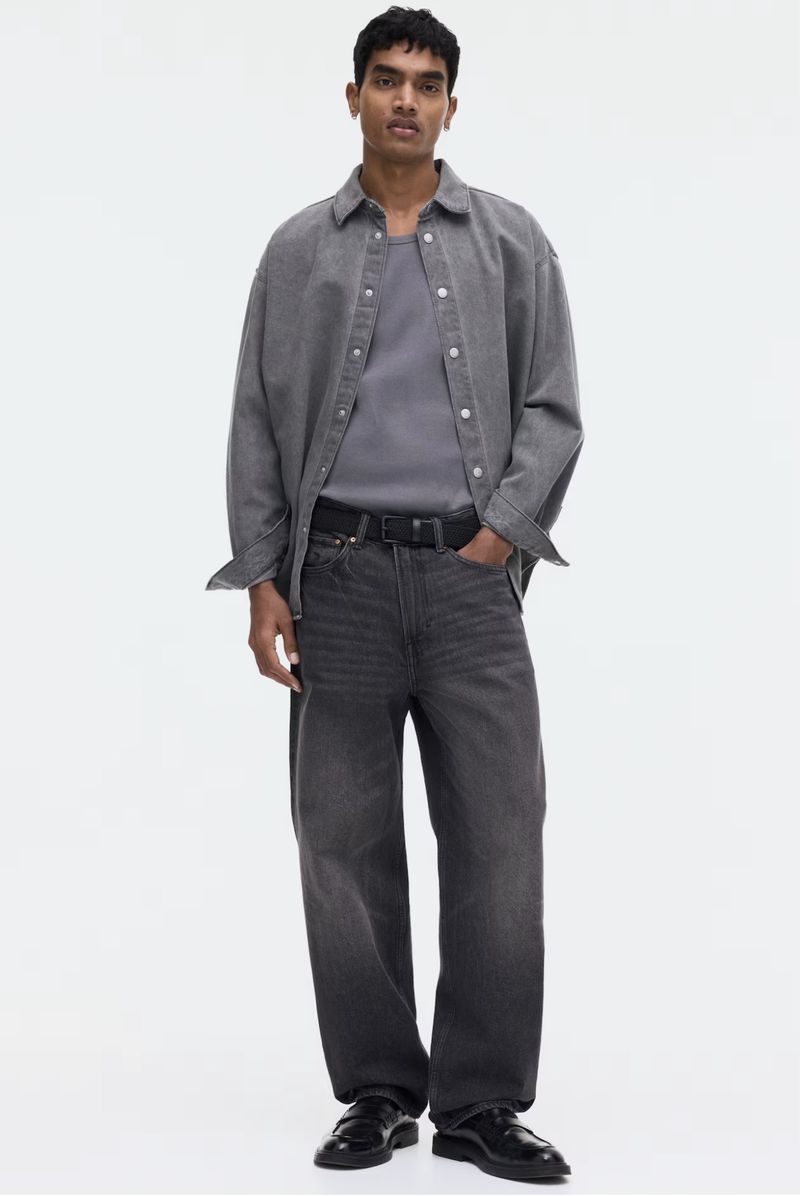 Full body studio shot of a young adult male wearing a gray shirt, light gray jacket, gray jeans, black belt, and black shoes.
