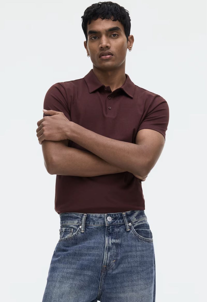 Man in burgundy polo shirt and blue jeans poses against a white backdrop with arms folded, fashion and portrait photography.