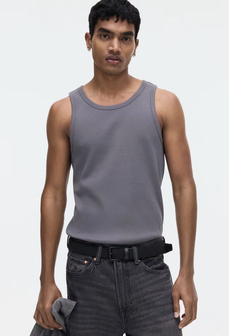 A young adult male poses indoors, wearing a gray tank top and faded black jeans with a black belt, set against a neutral background.