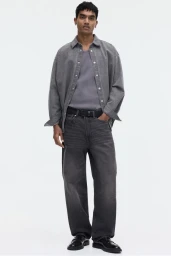 A man wearing an open grey overshirt, grey t-shirt, and dark jeans against a white background